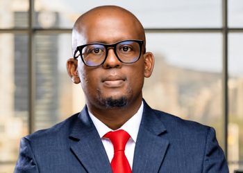 Portrait of a bald man in a navy suit and red tie, wearing black-ramed glasses, looking at the camera in an office setting