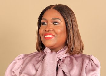 Smiling woman with shoulder-length brown hair wearing a pink blouse against a beige background.
