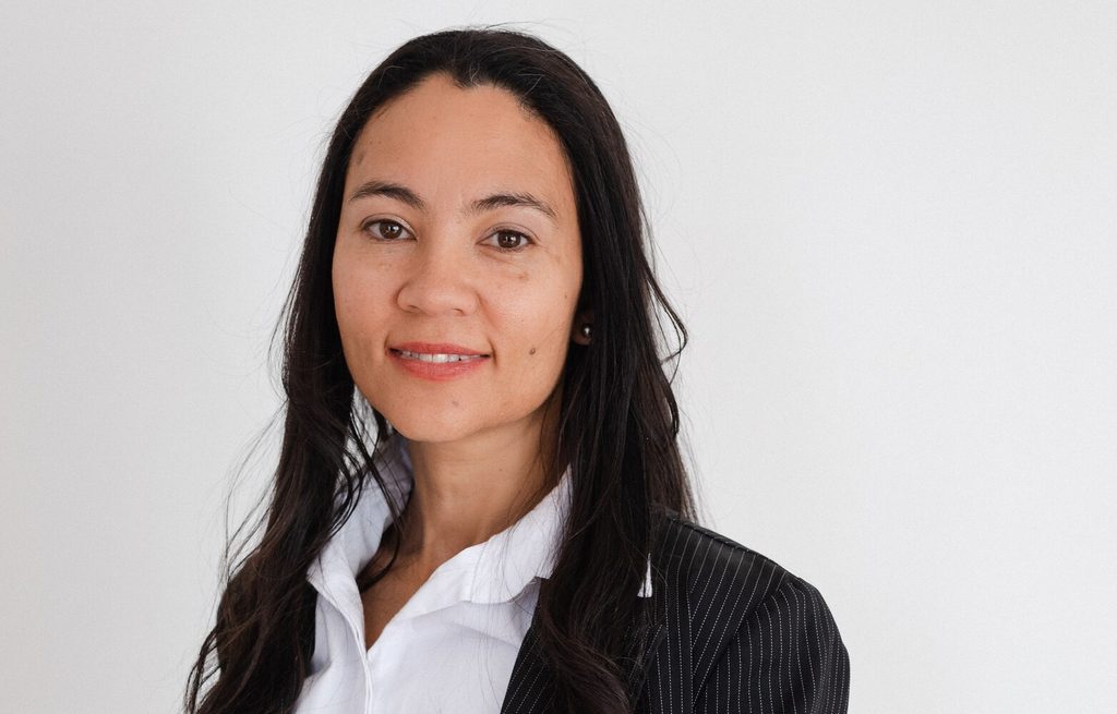 Professional headshot of a woman with long dark hair, wearing a white blouse and pinstripe blazer against a light background.