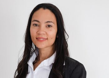Professional headshot of a woman with long dark hair, wearing a white blouse and pinstripe blazer against a light background.