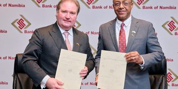 Two businessmen in suits pose with documents at a Bank of Namibia event backdrop.