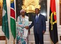 Two officials shake hands in a formal government setting, with a gold emblem behind them and multiple national flags on either side.