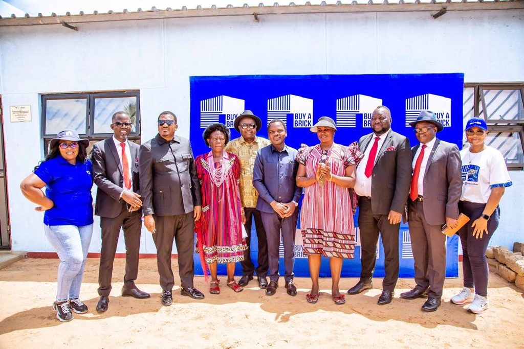 WhatsApp Image 2026-10-14 at 61108 PM - The Brief | Namibia's Leading Business & Financial News Ten people stand in a line outdoors, smiling, in front of a blue 'Buy a Brick' backdrop at a community event.