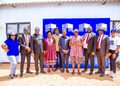 Ten people stand in a line outdoors, smiling, in front of a blue 'Buy a Brick' backdrop at a community event.