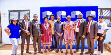Ten people stand in a line outdoors, smiling, in front of a blue 'Buy a Brick' backdrop at a community event.