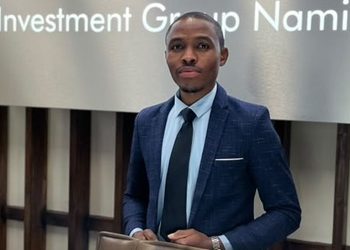 Professional man in a dark blue suit and light blue shirt stands in front of a wall sign reading 'Investment Group Namibia,' holding a tablet.