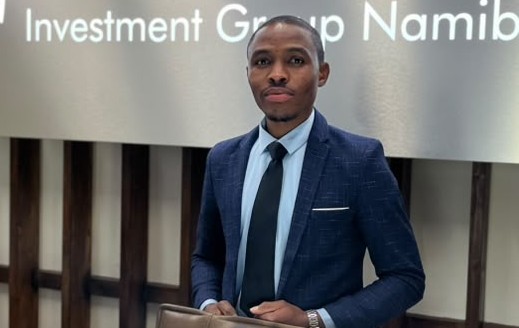 Titus Shigwedha - The Brief | Namibia's Leading Business & Financial News Professional man in a dark blue suit and light blue shirt stands in front of a wall sign reading 'Investment Group Namibia,' holding a tablet.