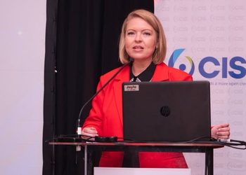 Karen Jansen van Vuuren - The Brief | Namibia's Leading Business & Financial News Woman in a red blazer delivering a talk at a podium with a laptop on stage at a conference.