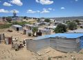 Hillside settlement with numerous corrugated metal shacks scattered across dry, dusty terrain under a bright blue sky with fluffy clouds.