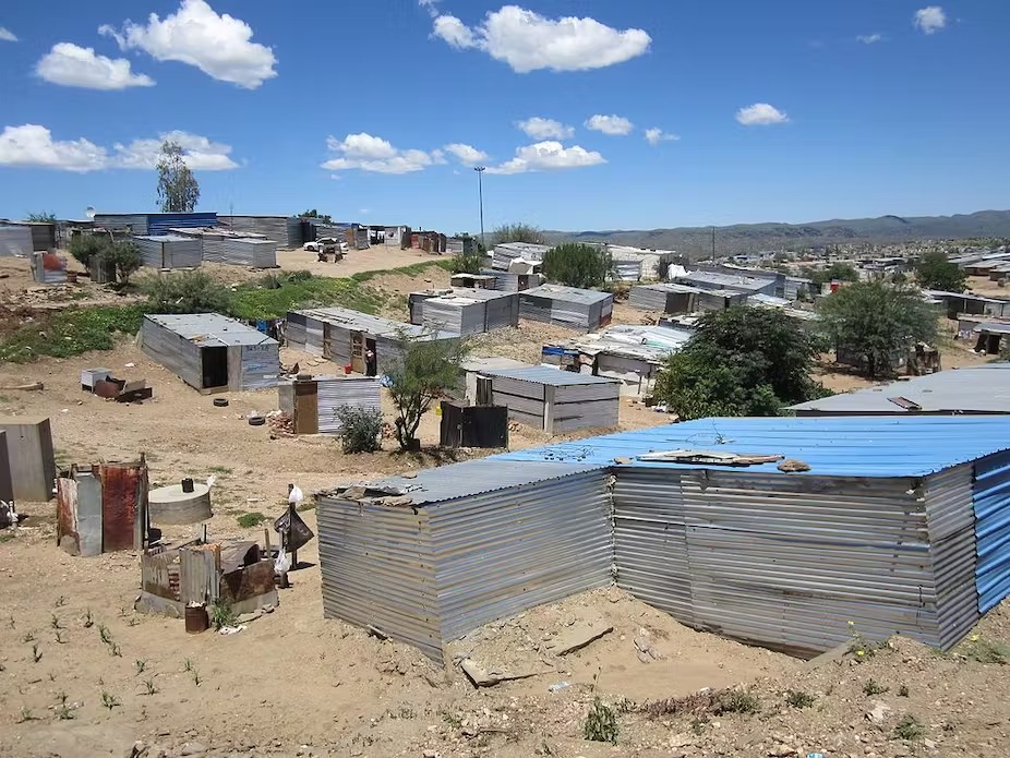 Hillside settlement with numerous corrugated metal shacks scattered across dry, dusty terrain under a bright blue sky with fluffy clouds.