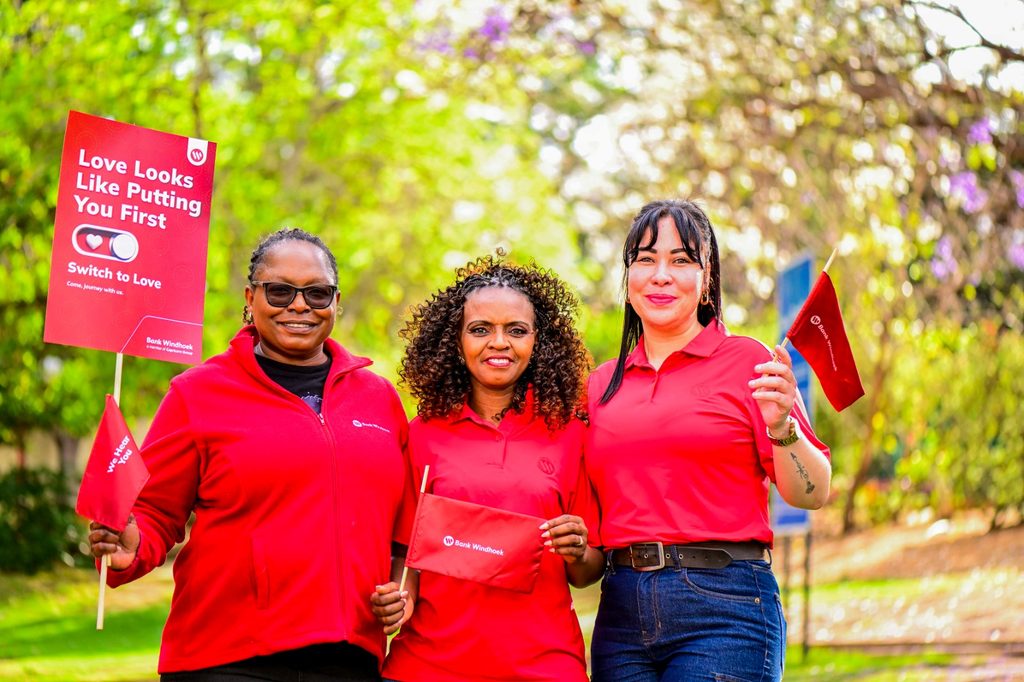 Three women in red shirts smile outdoors, holding red campaign banners and flags for a 'Love Looks Like Putting You First' event by Bank Windhoek.
