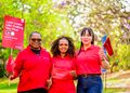 Three women in red shirts smile outdoors, holding red campaign banners and flags for a 'Love Looks Like Putting You First' event by Bank Windhoek.