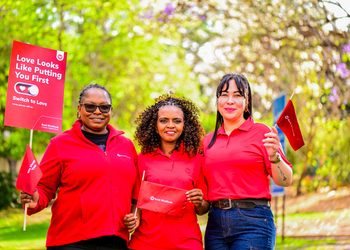 Three women in red shirts smile outdoors, holding red campaign banners and flags for a 'Love Looks Like Putting You First' event by Bank Windhoek.