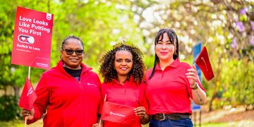 Three women in red shirts smile outdoors, holding red campaign banners and flags for a 'Love Looks Like Putting You First' event by Bank Windhoek.