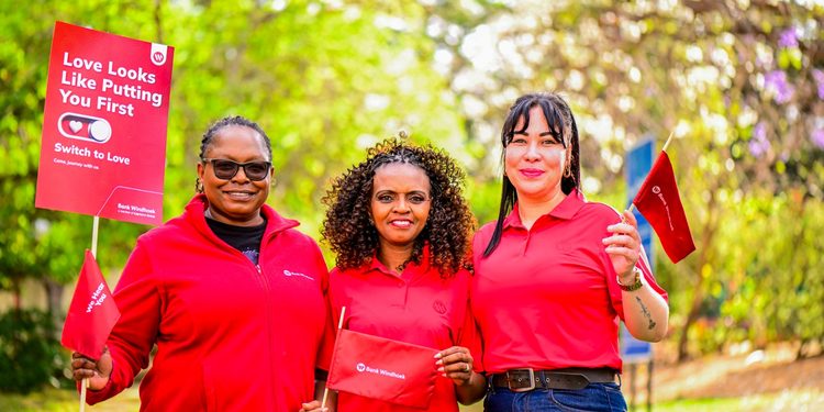 Three women in red shirts smile outdoors, holding red campaign banners and flags for a 'Love Looks Like Putting You First' event by Bank Windhoek.