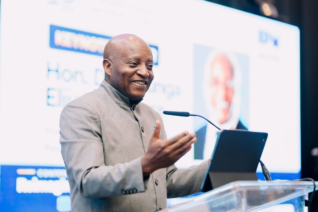 NGURARE0 - The Brief | Namibia's Leading Business & Financial News Smiling man in a gray suit gives a conference talk, gesturing with his hands beside a tablet on a clear podium.