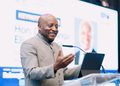 Smiling man in a gray suit gives a conference talk, gesturing with his hands beside a tablet on a clear podium.