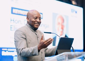 Smiling man in a gray suit gives a conference talk, gesturing with his hands beside a tablet on a clear podium.