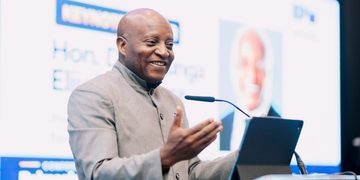 Smiling man in a gray suit gives a conference talk, gesturing with his hands beside a tablet on a clear podium.