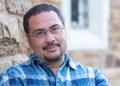 Man with short dark hair and glasses leaning against a stone wall outdoors, wearing a blue plaid shirt.