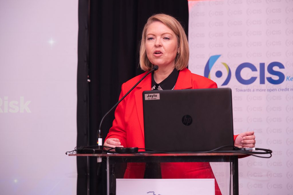 KARIN1 - The Brief | Namibia's Leading Business & Financial News Speaker in a red blazer standing behind a lectern with a laptop, microphone, and a visible name badge reading 'Jaylo' at a CIS-themed event.