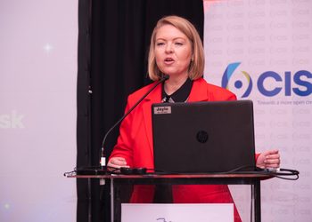 KARIN1 - The Brief | Namibia's Leading Business & Financial News Speaker in a red blazer standing behind a lectern with a laptop, microphone, and a visible name badge reading 'Jaylo' at a CIS-themed event.