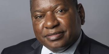Portrait of a middle-aged Black man in a dark suit and patterned tie against a gray background, looking at the camera.