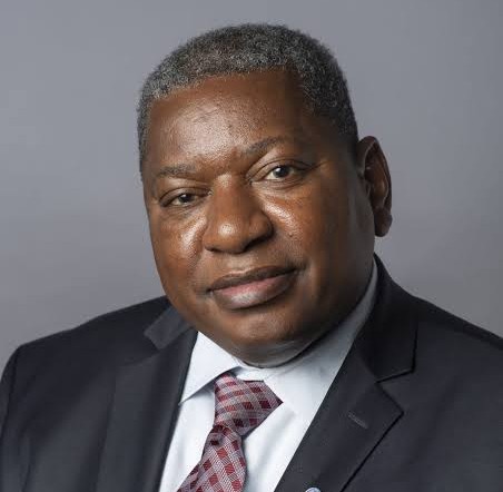 Portrait of a middle-aged Black man in a dark suit and patterned tie against a gray background, looking at the camera.