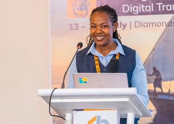 Woman speaker at a podium with a laptop and microphone, presenting at a conference with a 'Digital Transformation' banner in the background.