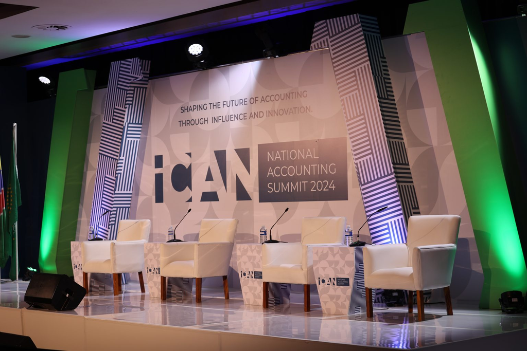 ICAN - The Brief | Namibia's Leading Business & Financial News Stage setup for ICAN National Accounting Summit 2024 with five white chairs, microphones, and water bottles on a patterned backdrop.