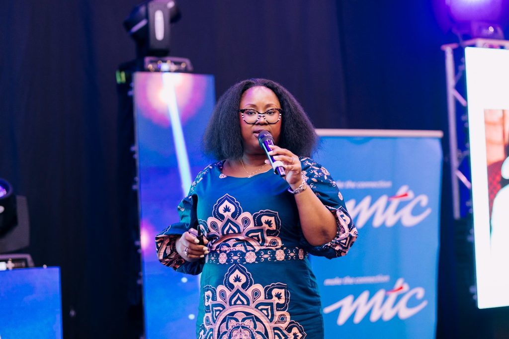 WhatsApp Image 2026-10-19 at 122531 - The Brief | Namibia's Leading Business & Financial News Black woman with glasses speaks into a handheld microphone on stage, wearing a teal patterned dress with a blue backdrop.