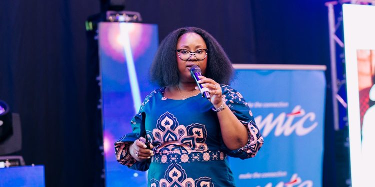 Black woman with glasses speaks into a handheld microphone on stage, wearing a teal patterned dress with a blue backdrop.