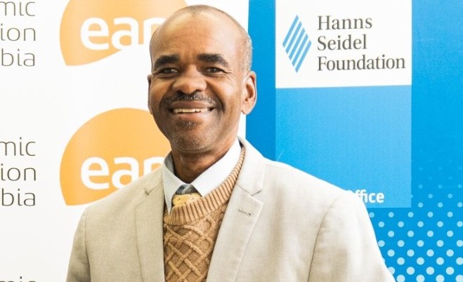 WhatsApp Image 2026-10-19 at 140652 - The Brief | Namibia's Leading Business & Financial News Smiling man in a beige blazer and tan sweater at a formal event, with banners for the Hanns Seidel Foundation in the background.