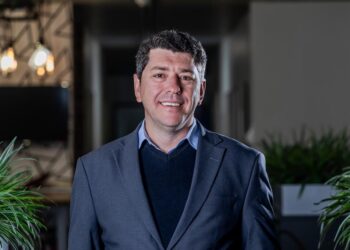 Portrait of a smiling man in a blue shirt and navy blazer in a modern office with plants nearby.