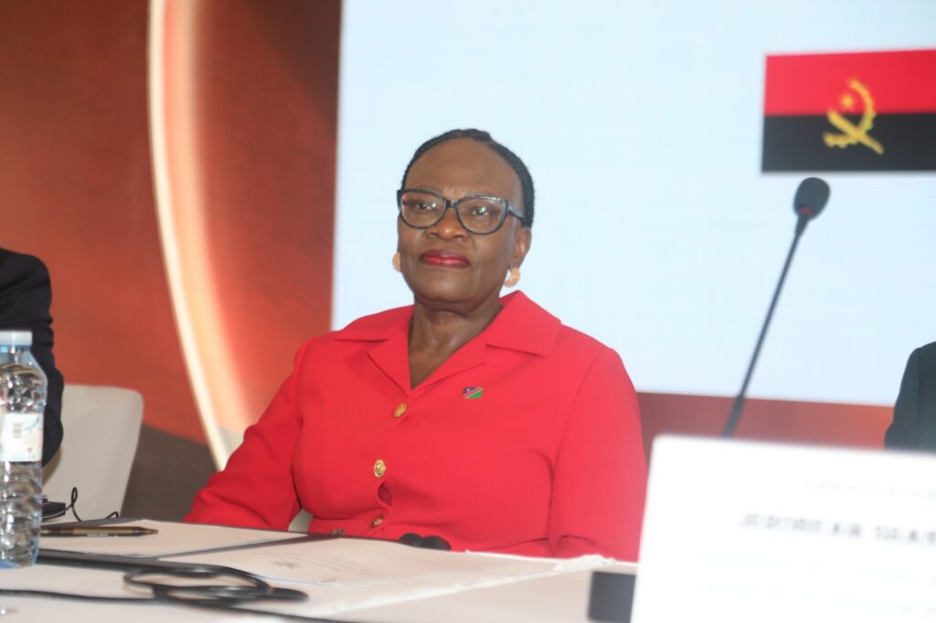 Panelist in a red blazer with glasses and gold earrings sits at a conference desk, microphone to her right and a small flag pin on her lapel.