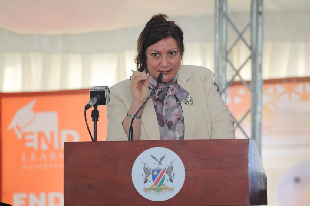 WhatsApp Image 2026-10-20 at 100122 - The Brief | Namibia's Leading Business & Financial News Woman in a beige blazer with a scarf speaks at a wooden lectern, delivering a speech into a microphone with a crest on the front panel.