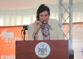 WhatsApp Image 2026-10-20 at 100122 - The Brief | Namibia's Leading Business & Financial News Woman in a beige blazer with a scarf speaks at a wooden lectern, delivering a speech into a microphone with a crest on the front panel.