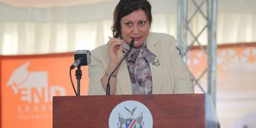 Woman in a beige blazer with a scarf speaks at a wooden lectern, delivering a speech into a microphone with a crest on the front panel.