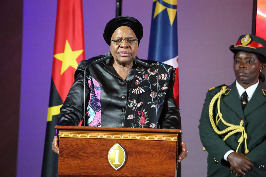 Woman in a black floral jacket speaking at a podium with a yellow emblem, flanked by flags and a uniformed officer on the right.