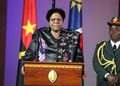 Woman in a black floral jacket speaking at a podium with a yellow emblem, flanked by flags and a uniformed officer on the right.