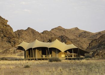tented lodge - The Brief | Namibia's Leading Business & Financial News Desert camp with a row of beige canvas tents set on a platform against rocky desert hills at dusk.