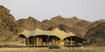 Desert camp with a row of beige canvas tents set on a platform against rocky desert hills at dusk.