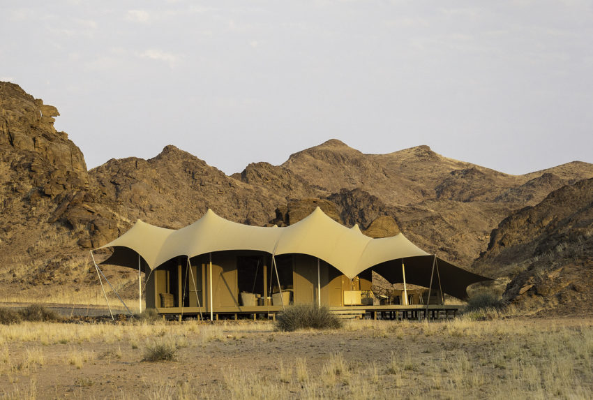 tented lodge - The Brief | Namibia's Leading Business & Financial News Desert camp with a row of beige canvas tents set on a platform against rocky desert hills at dusk.