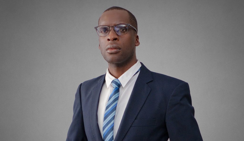 Dickson Matengu - The Brief | Namibia's Leading Business & Financial News Portrait of a man in a navy suit and blue striped tie, wearing glasses, against a gray backdrop.