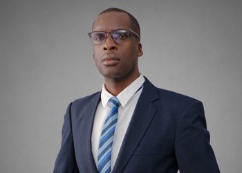 Dickson Matengu - The Brief | Namibia's Leading Business & Financial News Portrait of a man in a navy suit and blue striped tie, wearing glasses, against a gray backdrop.