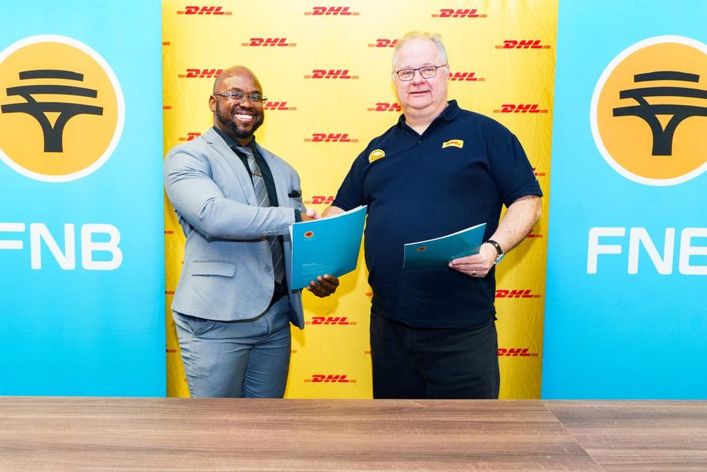 WhatsApp Image 2026-10-20 at 104716 PM - The Brief | Namibia's Leading Business & Financial News Two men shake hands after signing an agreement; one in a gray suit, the other in a dark polo, in front of DHL banners with FNB logos nearby.