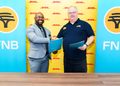 Two men shake hands after signing an agreement; one in a gray suit, the other in a dark polo, in front of DHL banners with FNB logos nearby.