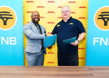WhatsApp Image 2026-10-20 at 104716 PM - The Brief | Namibia's Leading Business & Financial News Two men shake hands after signing an agreement; one in a gray suit, the other in a dark polo, in front of DHL banners with FNB logos nearby.