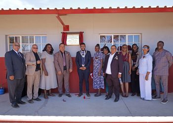 Group of formally dressed adults posing outside a school for a ribbon-cutting/inauguration ceremony by a plaque on the wall with red curtain backdrop.