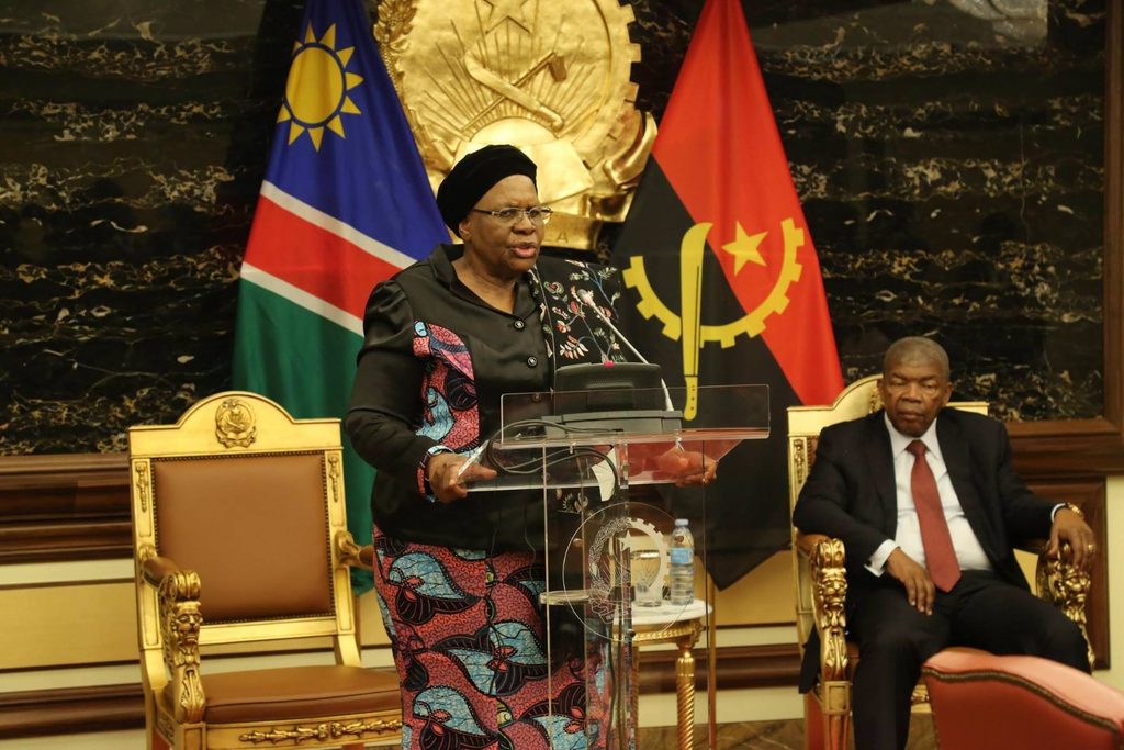 Woman in a patterned dress speaks at a clear podium with microphone, flanked by Namibian flags and a seated man in a suit behind her in a formal government setting